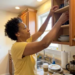 Angela reaching both arms overhead into a kitchen cabinet, no wince